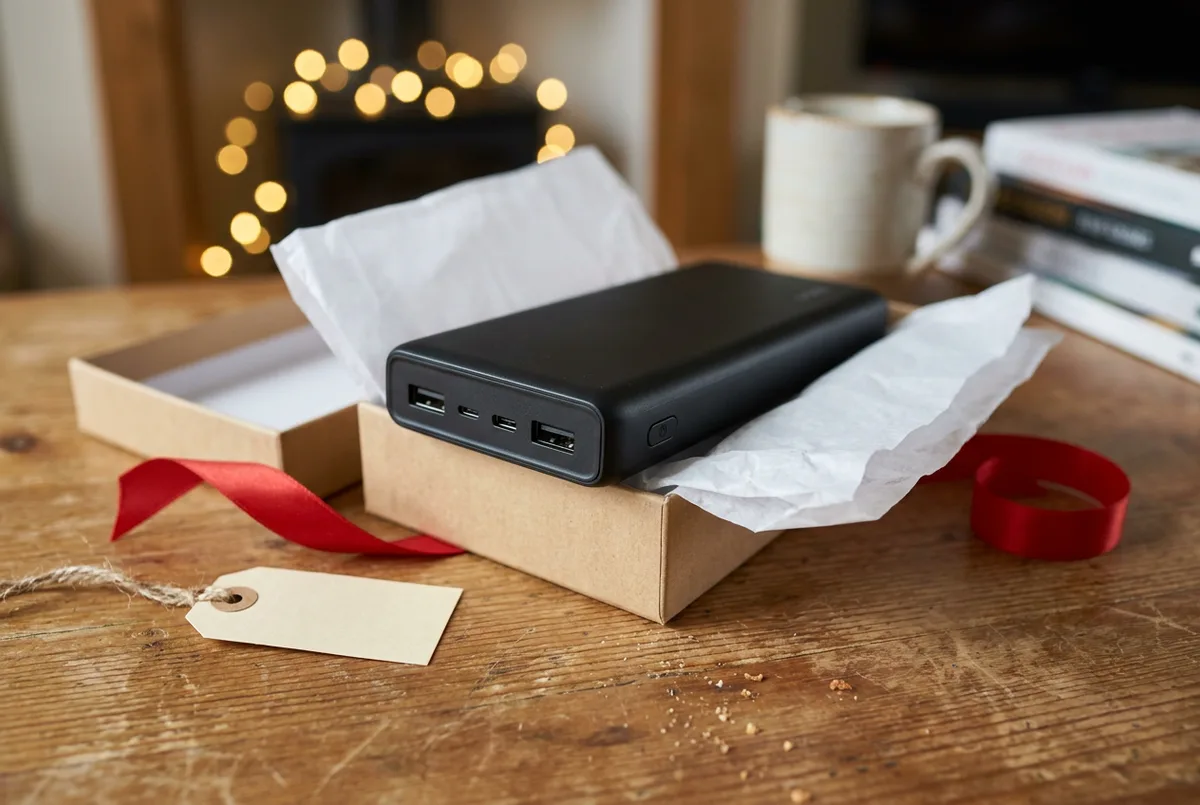 A dark gray portable power bank charging a smartphone on a light wood nightstand.